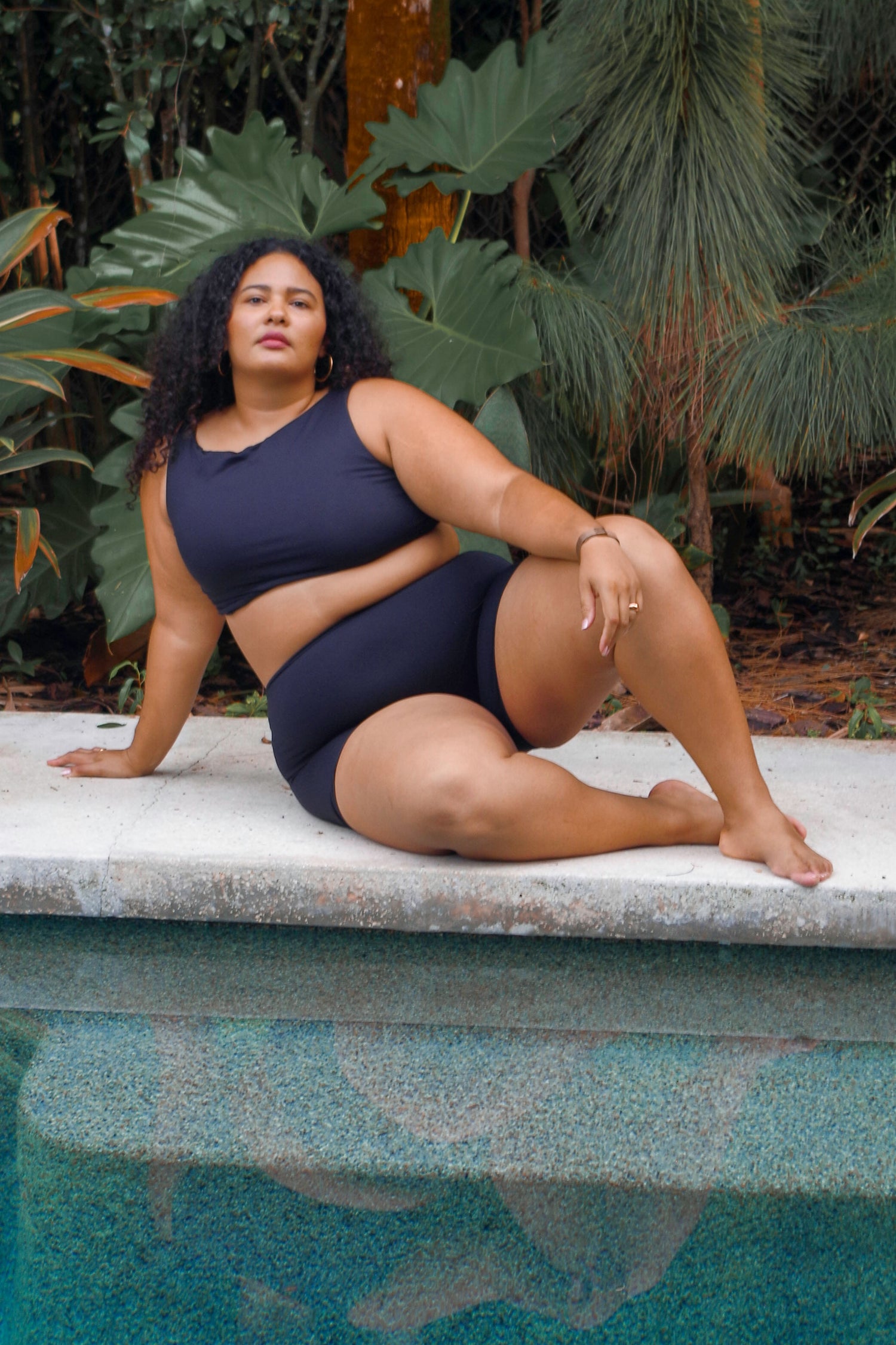 plus size woman wearing cropped black tankini with removable padding and high waisted swim shorts laying by swimming pool